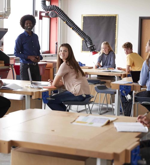 Class Of High School Students Sitting At Work Benches Listening To Teacher In Design And Technology Lesson
