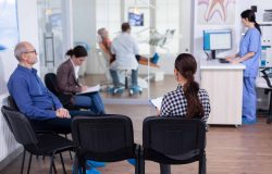 Crowded stomatology waiting area with people filling form for dental consultation. Stomatoloy specialist denstiry treating senior woman cavity. Receptionist working on computer.
