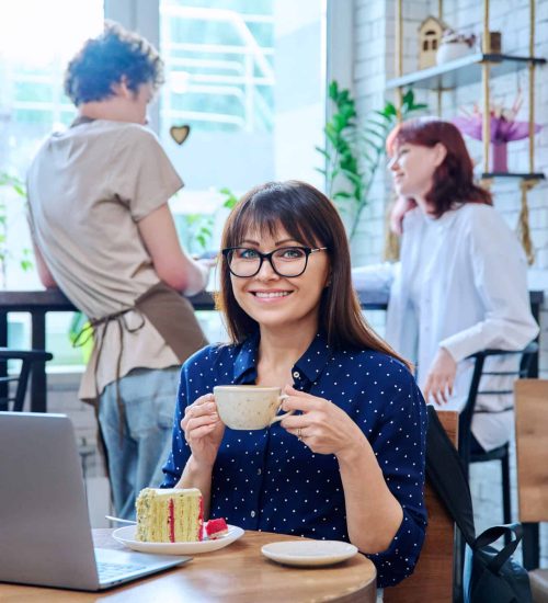Middle aged woman in bakery cafeteria with cup of coffee and dessert cake, sitting at table, with laptop, happy smiling female looking at camera. Lunch break, food, lifestyle, mature 40s people concept