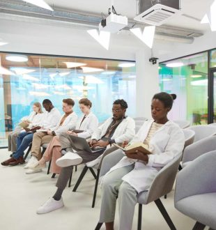 Vertical full length portrait of young African American woman wearing lab coat while sitting in row with multi-ethnic group of people in audience at medical seminar, copy space