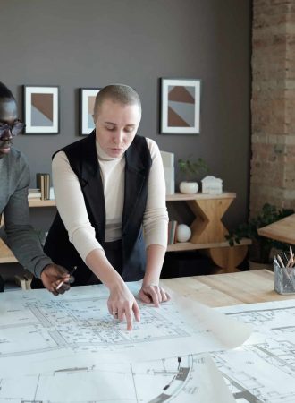 Two confident engineers in smart casualwear discussing sketch on blueprint while standing by table in large office