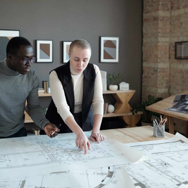 Two confident engineers in smart casualwear discussing sketch on blueprint while standing by table in large office