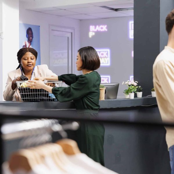 Black Friday and retail industry. Young Asian woman customer standing at clothing store checkout counter giving clothes to friendly cashier, buying garment at discounted prices during seasonal sales